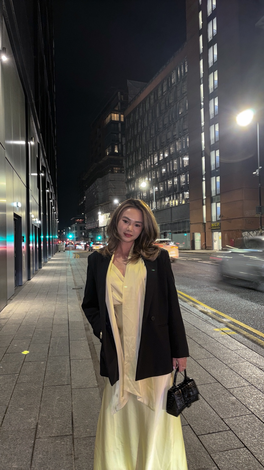 Candace wearing a pale yellow dress and black jacket on a city street at night